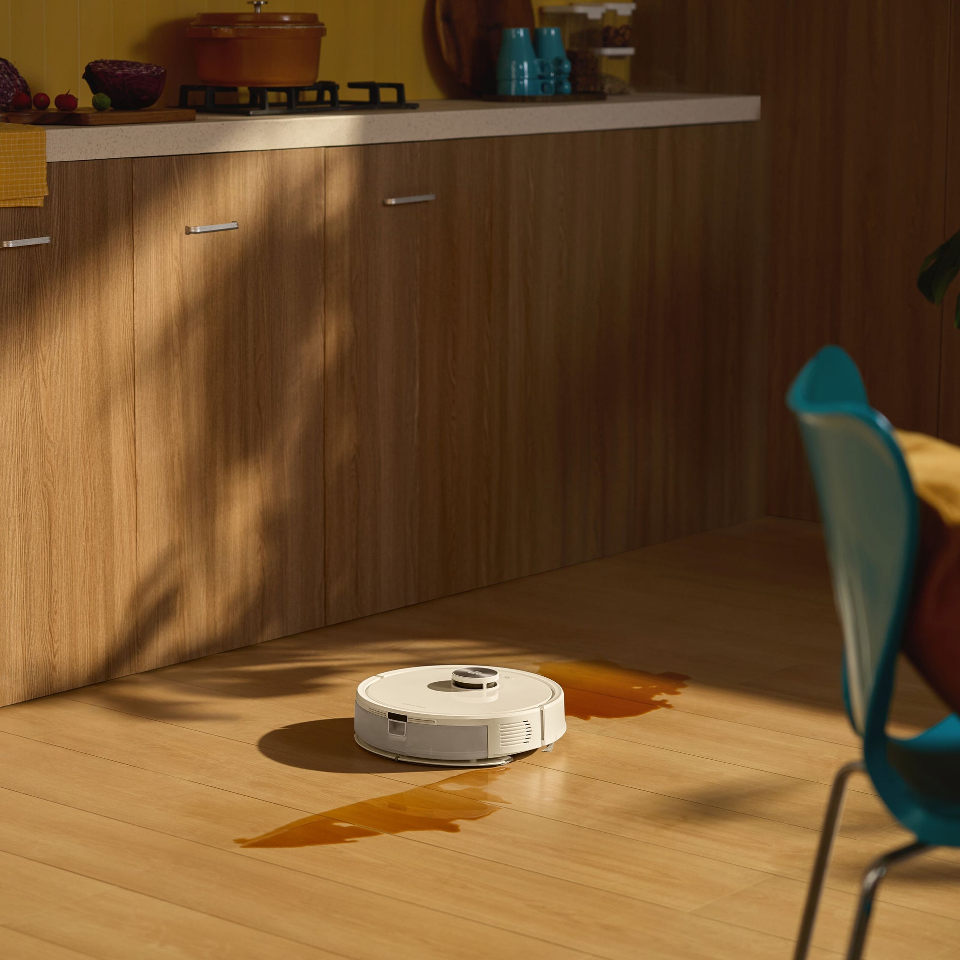 Roborock Q7 TF+ robotstøvsuger med ladestation fjerner orange væskespild på trægulv ved træskabe og blå stol.