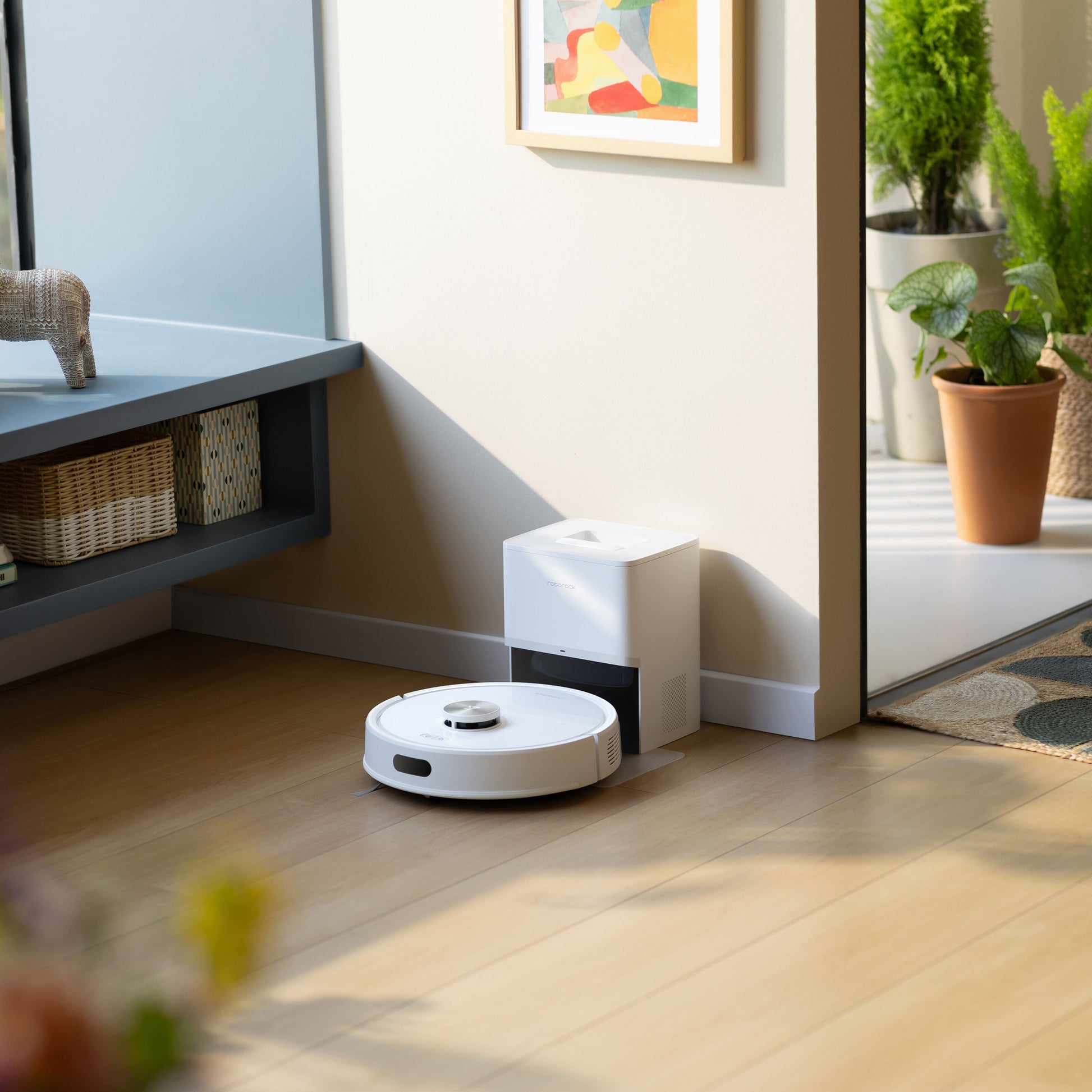Roborock Q7 TF+ robotstøvsuger med ladestation i hvid ved base på lyst trægulv med planter og abstrakt vægkunst.