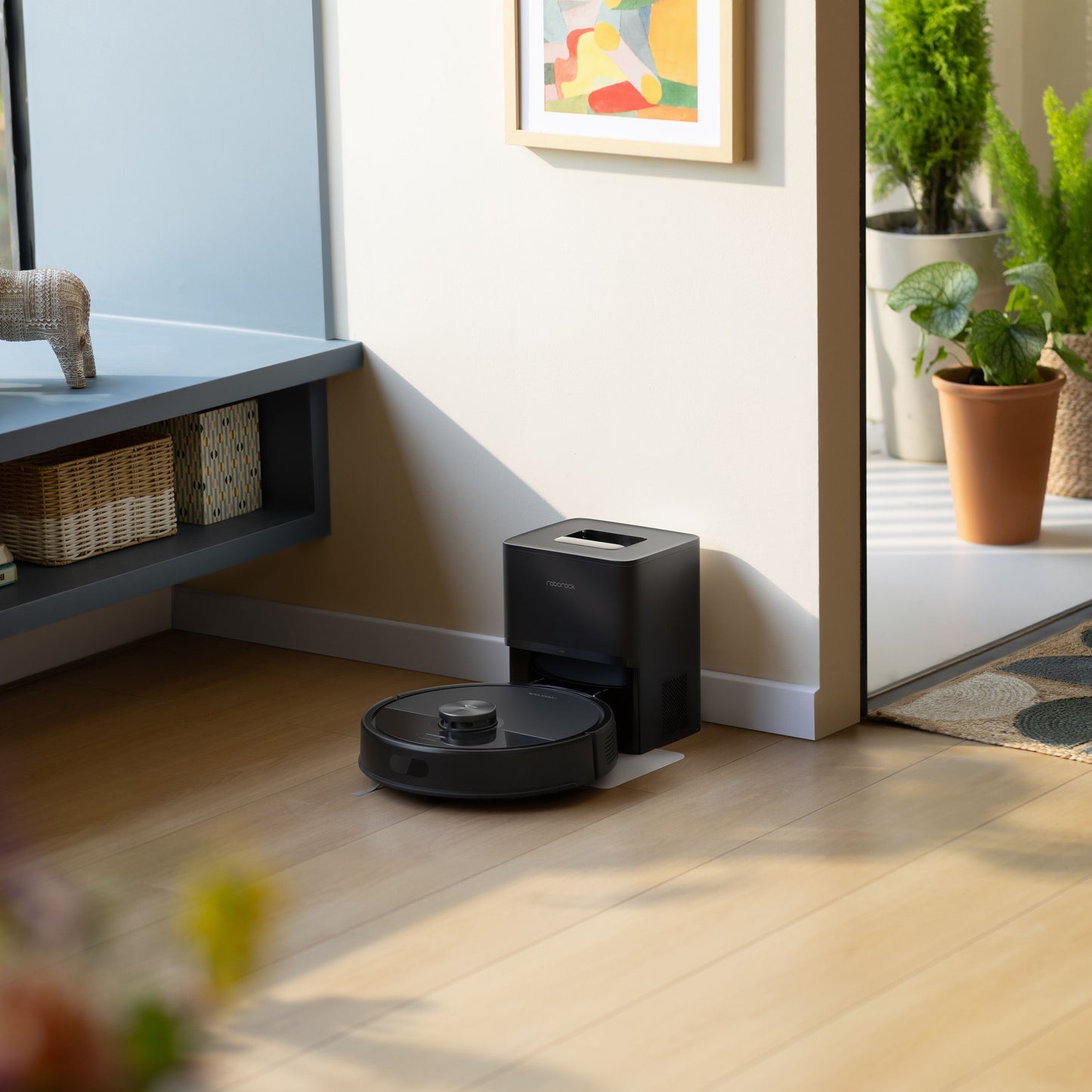 Roborock Q7 TF+ robotstøvsuger sorterer med ladestation på lyst trægulv, synlige potteplanter og fletkurv.