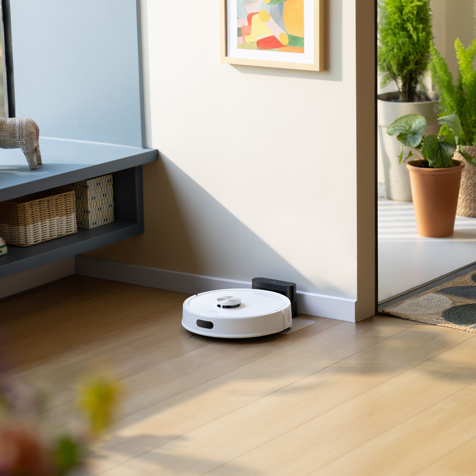 Roborock Q7 TF robotstøvsuger hvid ved ladestation i rum med trægulv, potteplante og abstrakt kunst.