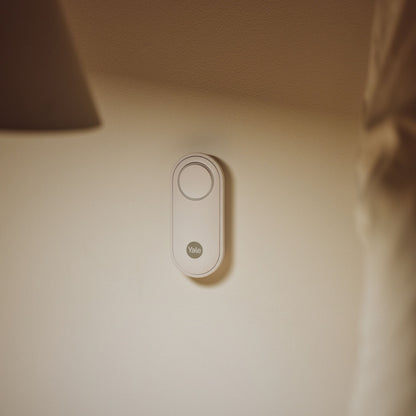 Yale Alarmknap oval monteret på lys væg, lampe og delvist synligt gardin i baggrunden.
