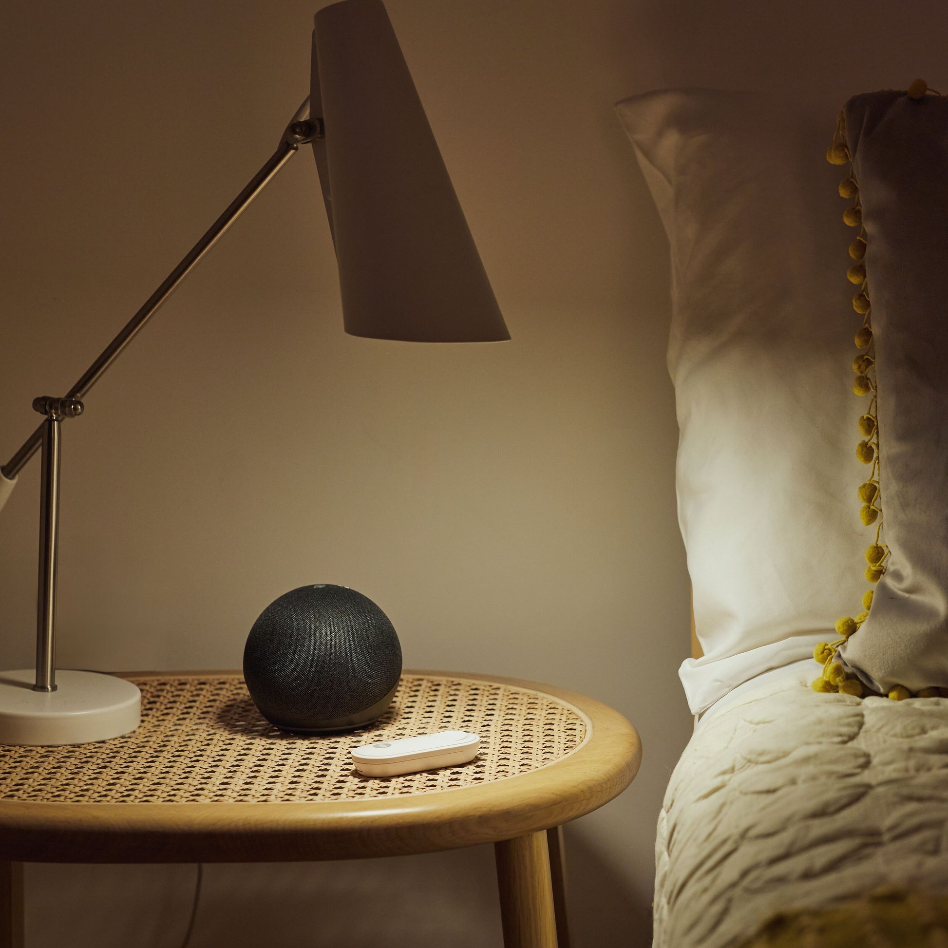 Sort smarthøjttaler, hvid fjernbetjening og lampe på rattan natbord, Yale Alarmknap med logo, seng med hvide puder og creme sengetæppe.