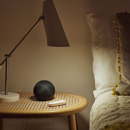 Sort smarthøjttaler, hvid fjernbetjening og lampe på rattan natbord, Yale Alarmknap med logo, seng med hvide puder og creme sengetæppe.