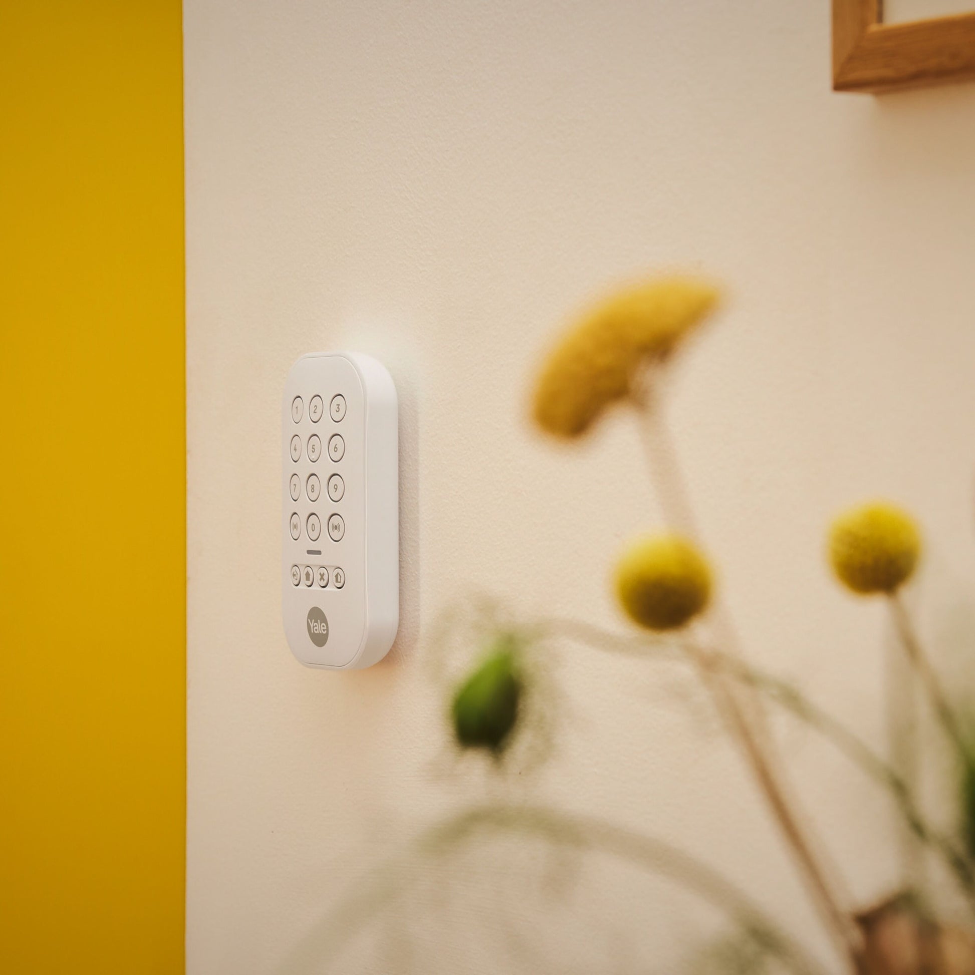 Yale Alarmknap sæt monteret på beige væg ved gul sektion, sløret gule blomster og træramme synlig i hjørnet.