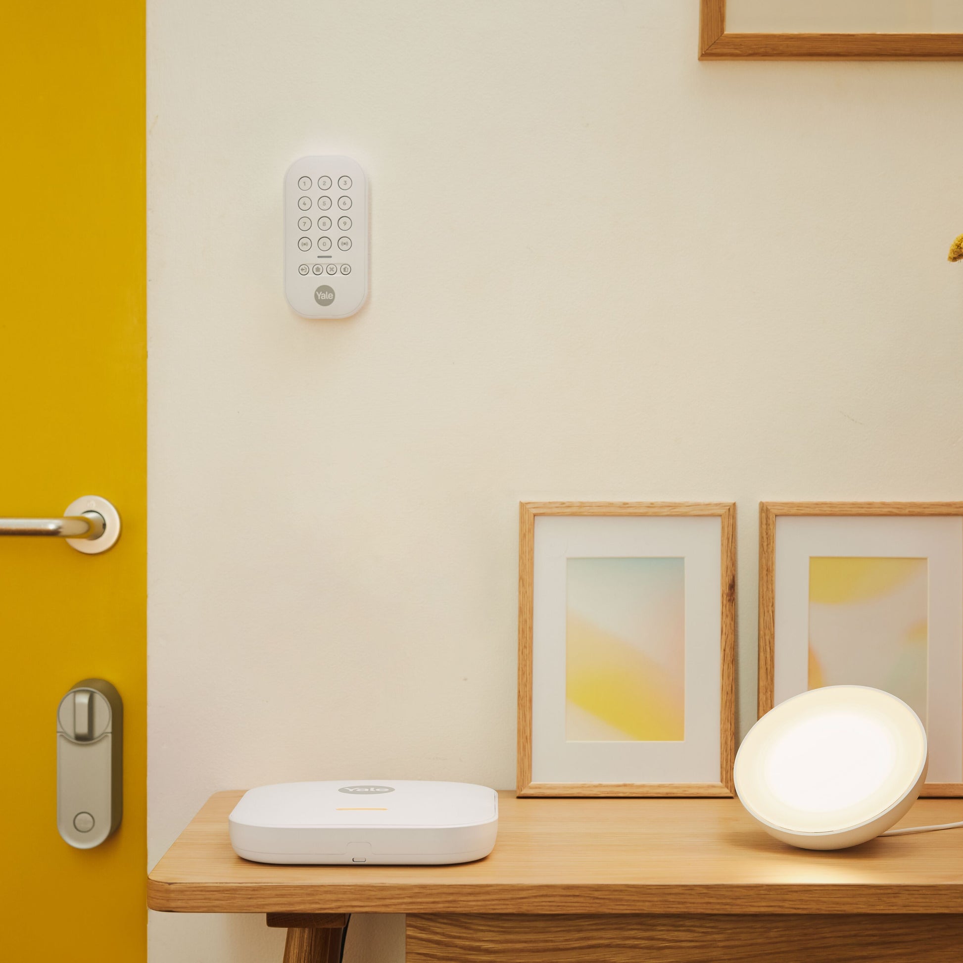 Yale Startalarmkit+ smartlåsesæt på gul dør med keypad, rund smart light og hvid enhed ved siden af på bord.