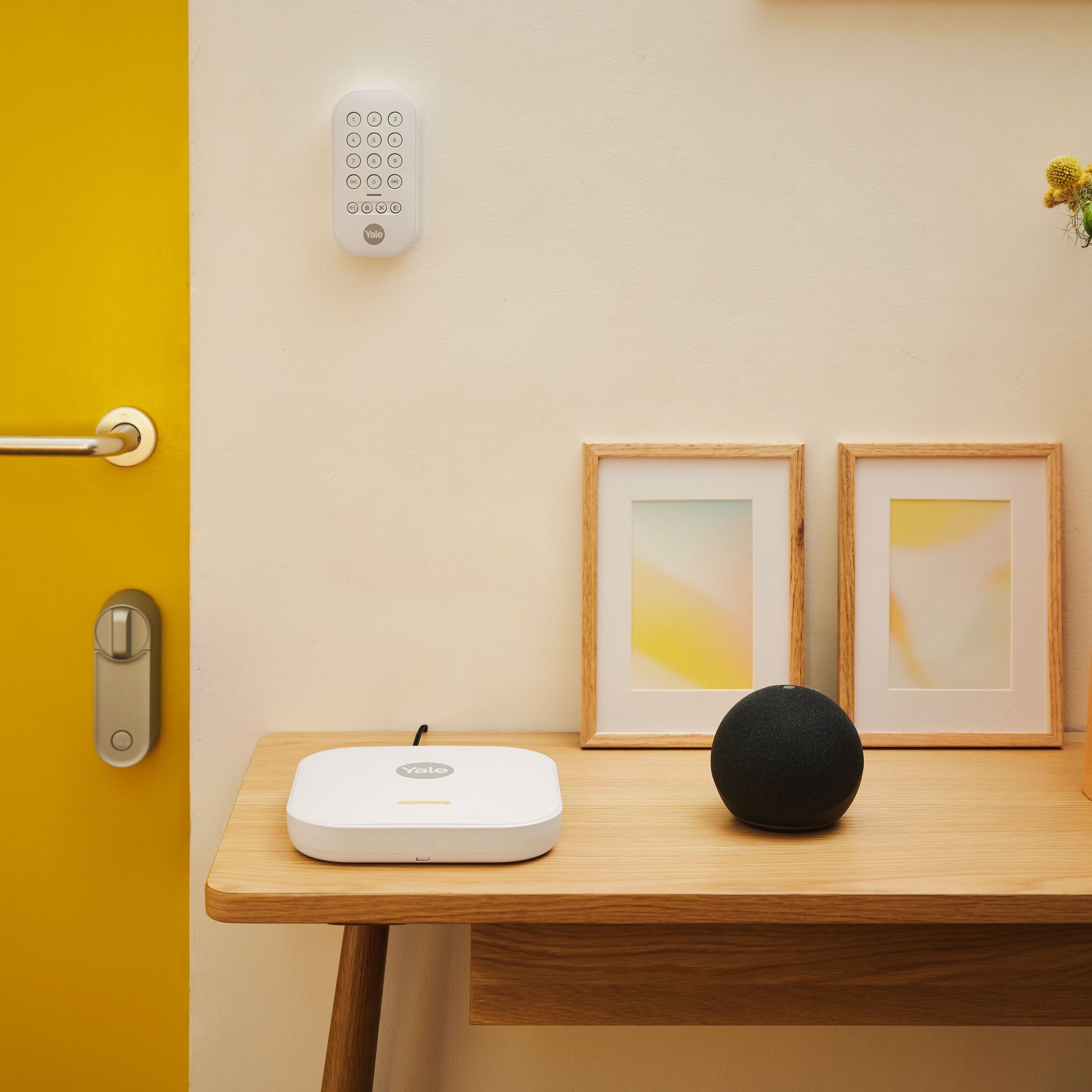 Yale Startalarmkit+ smartlås og tastatur på gul dør, hvid hub og sort smart højttaler på bord.
