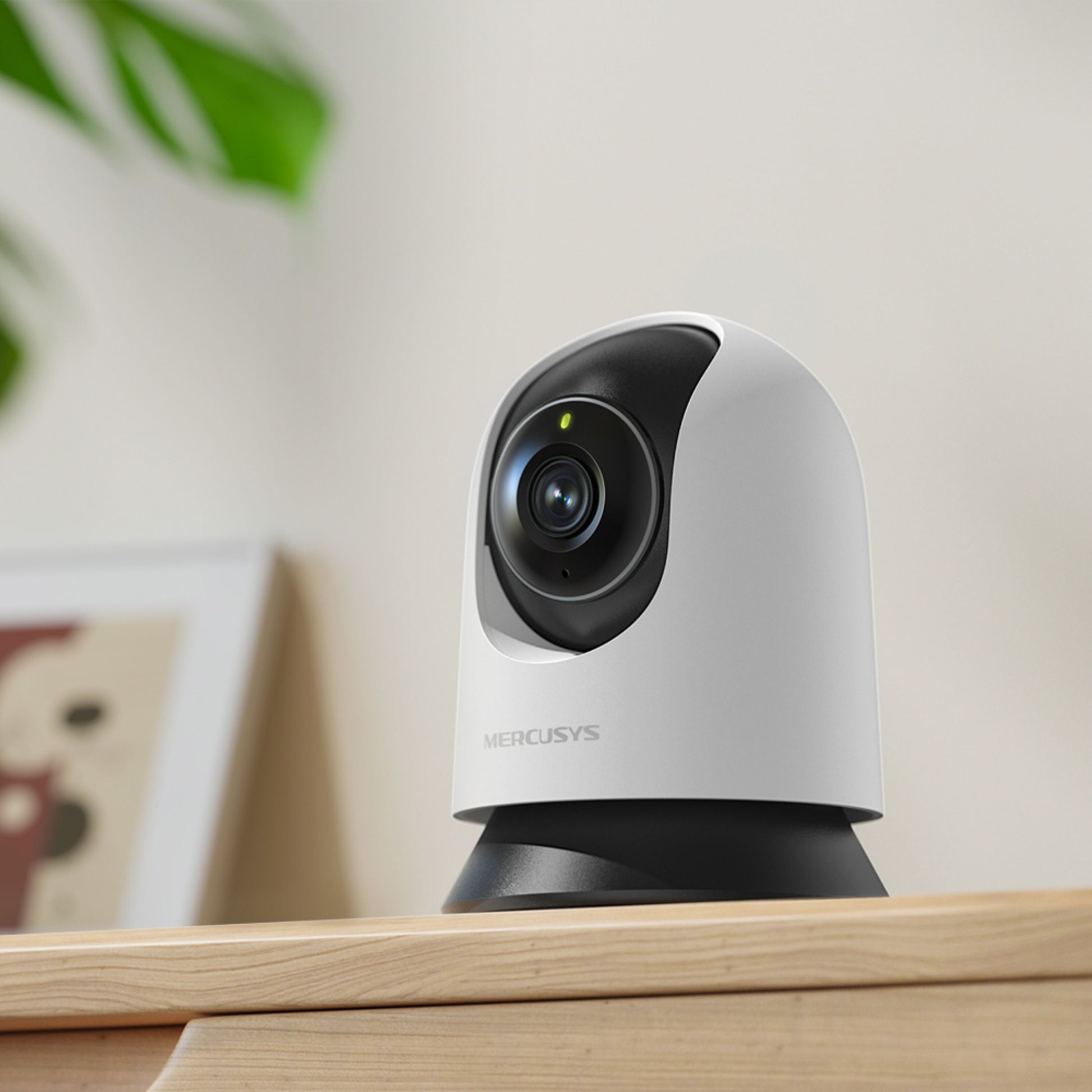Mercusys MC210 pan/tilt Wi-Fi overvågningskamera, hvid, på træoverflade med sløret plante og billedramme i baggrunden.