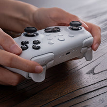 8Bitdo Ultimate 2 Bluetooth Controller hvid med sorte knapper, sat i hænder på træoverflade.