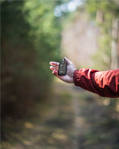Sort GPS-enhed og Goobay Powerbank til Apple Watch i hånd, bæres af person med rød jakke, skovbaggrund sløret.