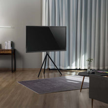 Fladskærms-TV på TV-gulvstander til 65", gråt tæppe på trægulv, lille bord med vase, grå lænestol, hvide gardiner.