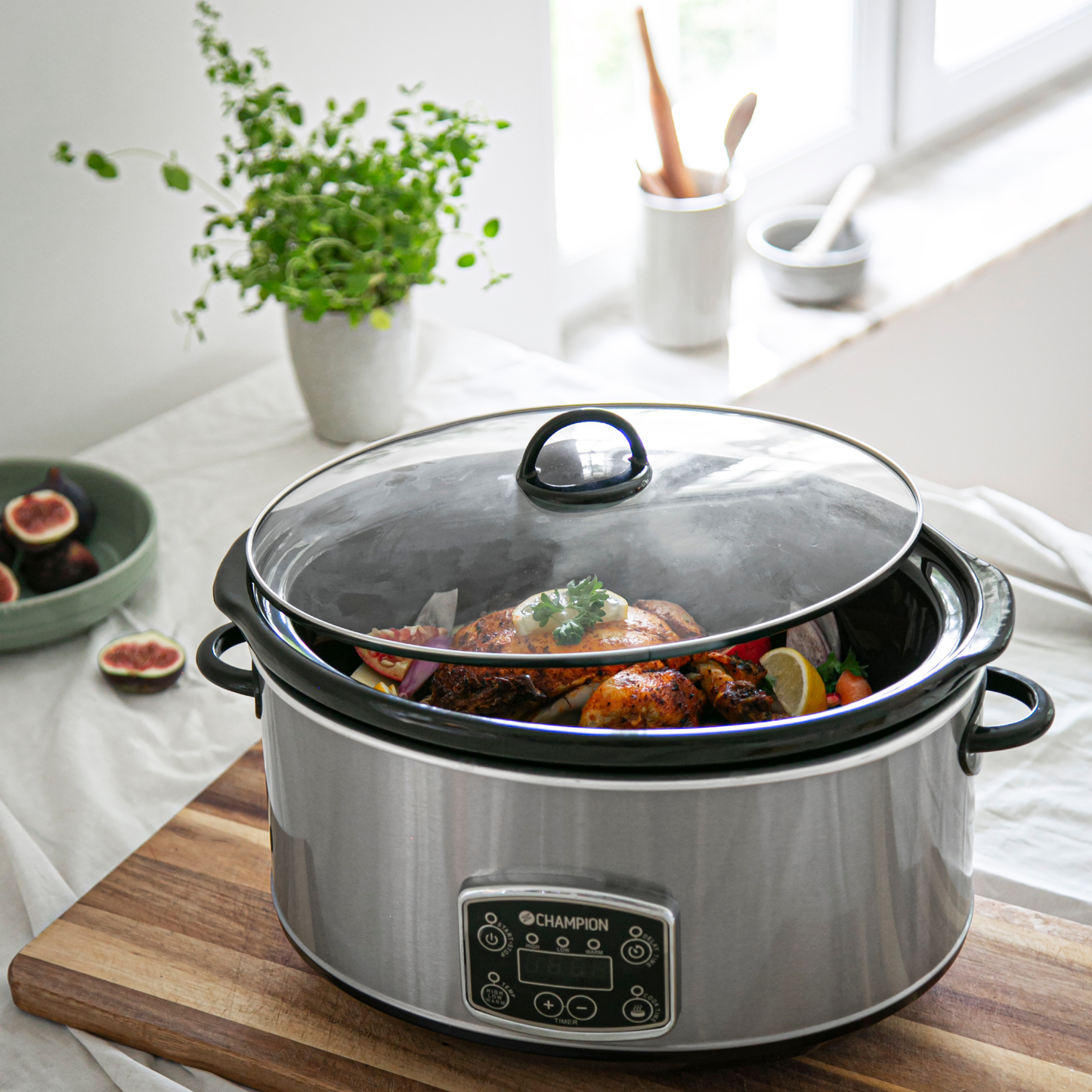 Champion slowcooker 6,5 L med stegt kylling og grøntsager i gryden, omgivet af friske krydderurter og figner.