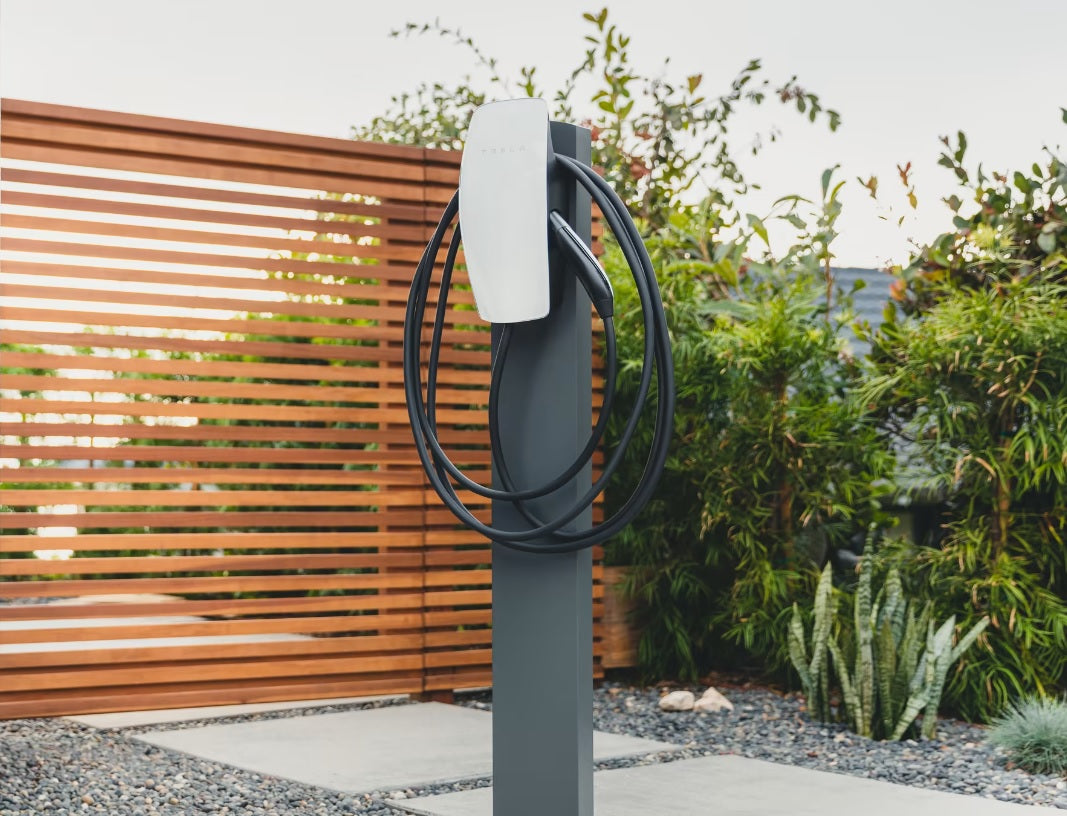 Tesla Wall Connector 22 kW hvid med spiral-ladekabel på stenflise, grønne planter og træhegn i baggrunden.