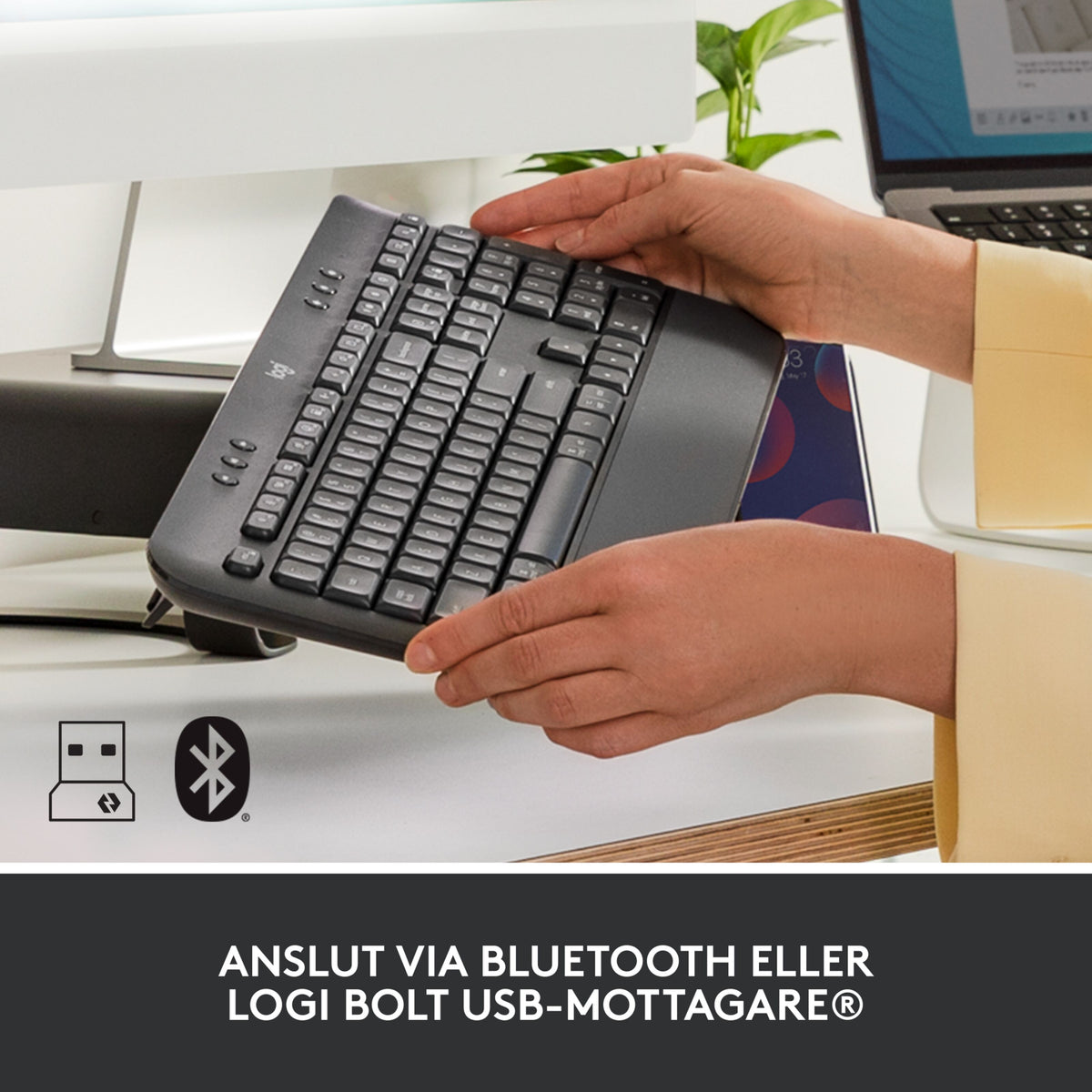 Hænder, der holder et grafitfarvet Logitech Signature K650 trådløst tastatur med håndledsstøtte og Bluetooth-/USB-ikoner.