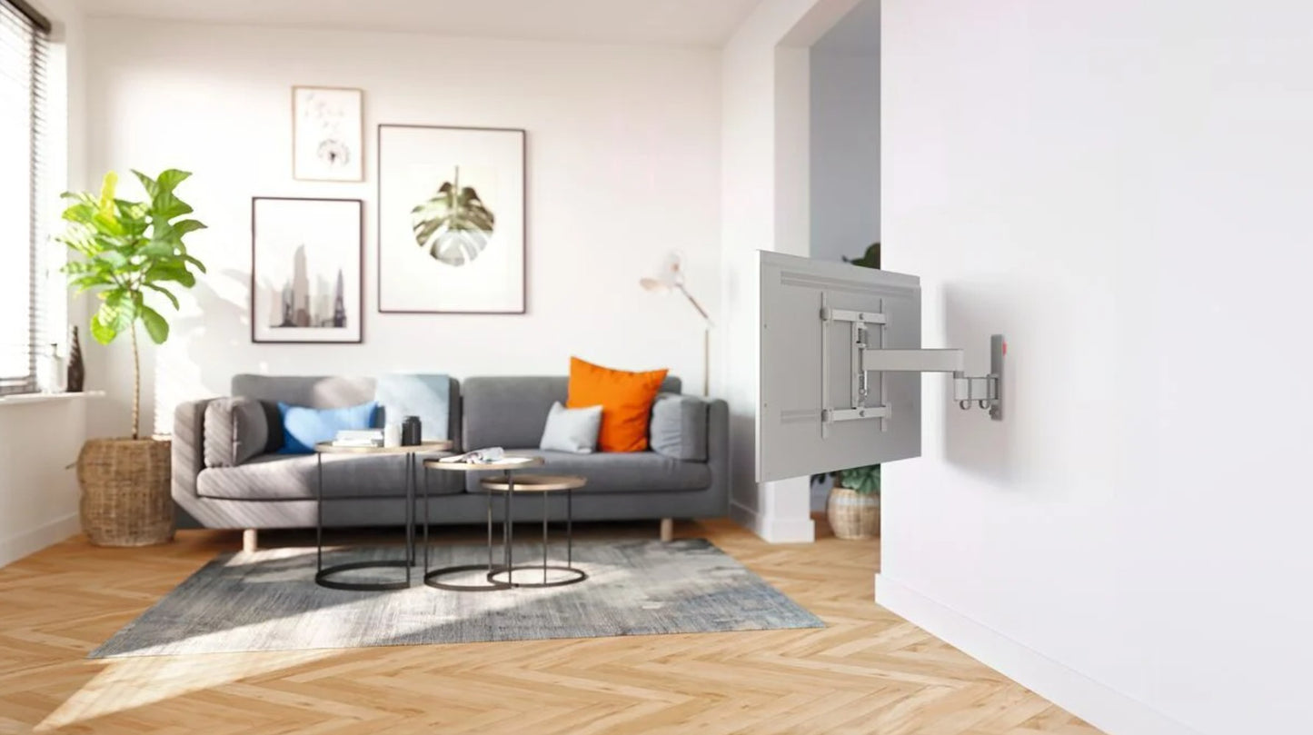 Vogels Comfort TVM 3445 32-65" vægbeslag med TV delvist drejet mod grå sofa, farvede puder, vægkunst, høj plante.