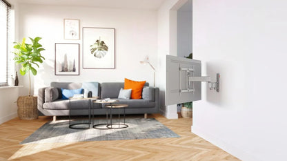 Vogels Comfort TVM 3445 32-65" vægbeslag med TV delvist drejet mod grå sofa, farvede puder, vægkunst, høj plante.