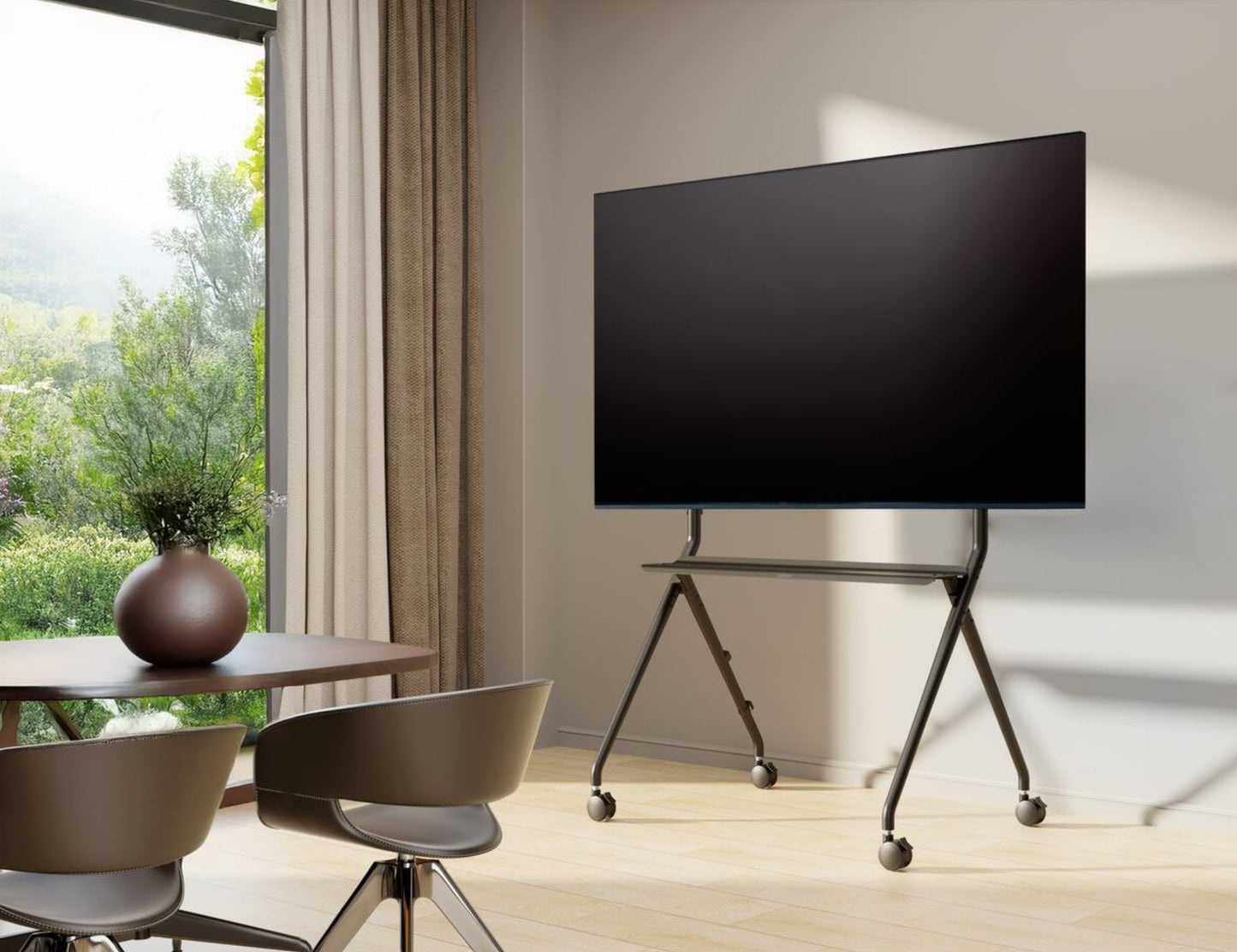 Vogels Essential T1011 B sort TV-gulvstativ med hjul, stor fladskærm monteret, to stole og rundt bord ved siden af.