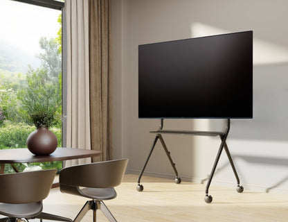 Vogels Essential T1011 B sort TV-gulvstativ med hjul, stor fladskærm monteret, to stole og rundt bord ved siden af.