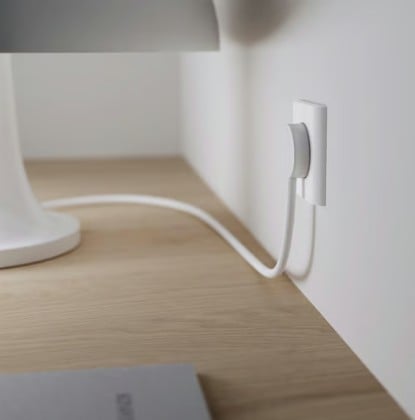 NORM K stikprop hvid uden jord tilsluttet hvidt vægstik på træbord, ledning synlig, sløret notesbog i forgrund.