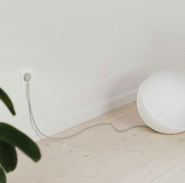 Hvid kuglelampe med NORM K stikprop grå uden jord i vægudtag på lyst trægulv nær hvid væg, grøn plante synlig.
