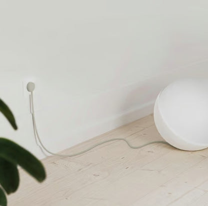 Hvid kuglelampe med NORM K stikprop grå uden jord i vægudtag på lyst trægulv nær hvid væg, grøn plante synlig.