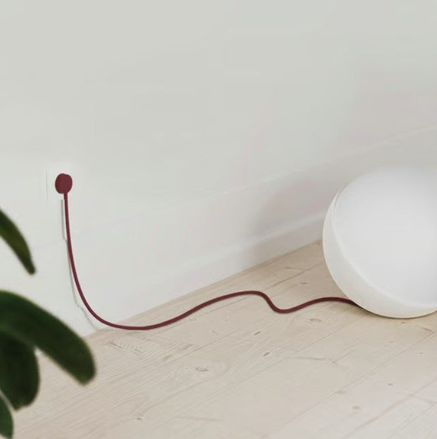 Hvid rund lampe med lang rød NORM K stikprop Rød Uden jord tilsluttet stikkontakt på lyst trægulv, grøn plante synlig.