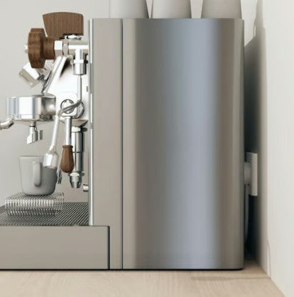 Stål espressomaskine med NORM K stikprop hvid med jord, kopper ovnpå, glas under portafilter på lys træbordplade.