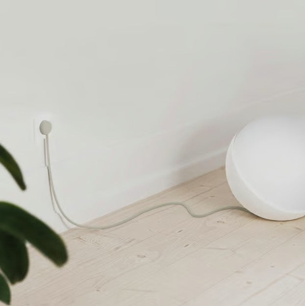 Rund hvid lampe med NORM K stikprop grå med jord, synlig ledning til stikkontakt, lyst trægulv, grøn plante i hjørne.