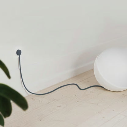 Rund hvid lampe med lang sort ledning tilsluttet NORM K stikprop blå med jord på lyst trægulv nær væg, grøn plante ses.
