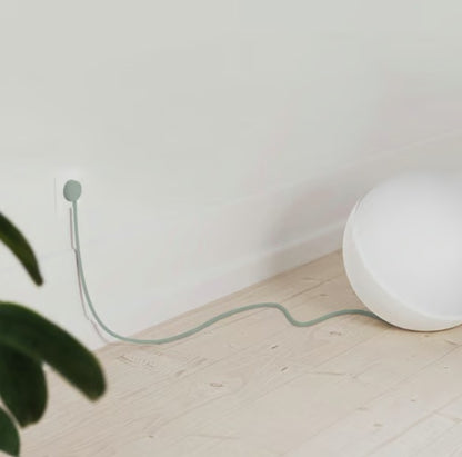 Rund hvid lampe med NORM K stikprop grøn med jord, tilsluttet stikkontakt på lyst trægulv ved hvid væg, grøn plante synlig.