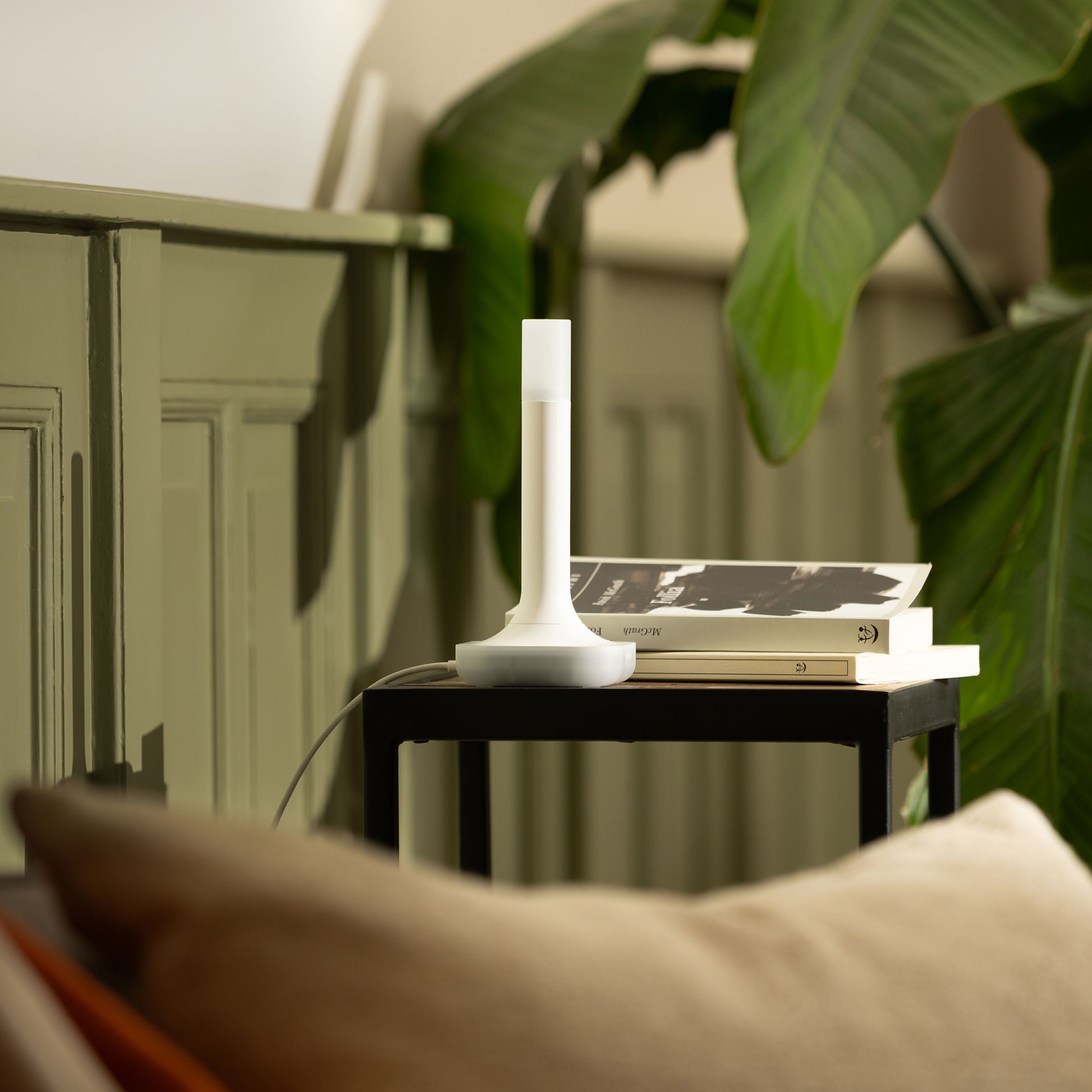 Hvid Zigbee 3.0 bordlampe model ZBT-2 på sort sidebord ved bøger, grøn væg og planter, beige sofa forrest.