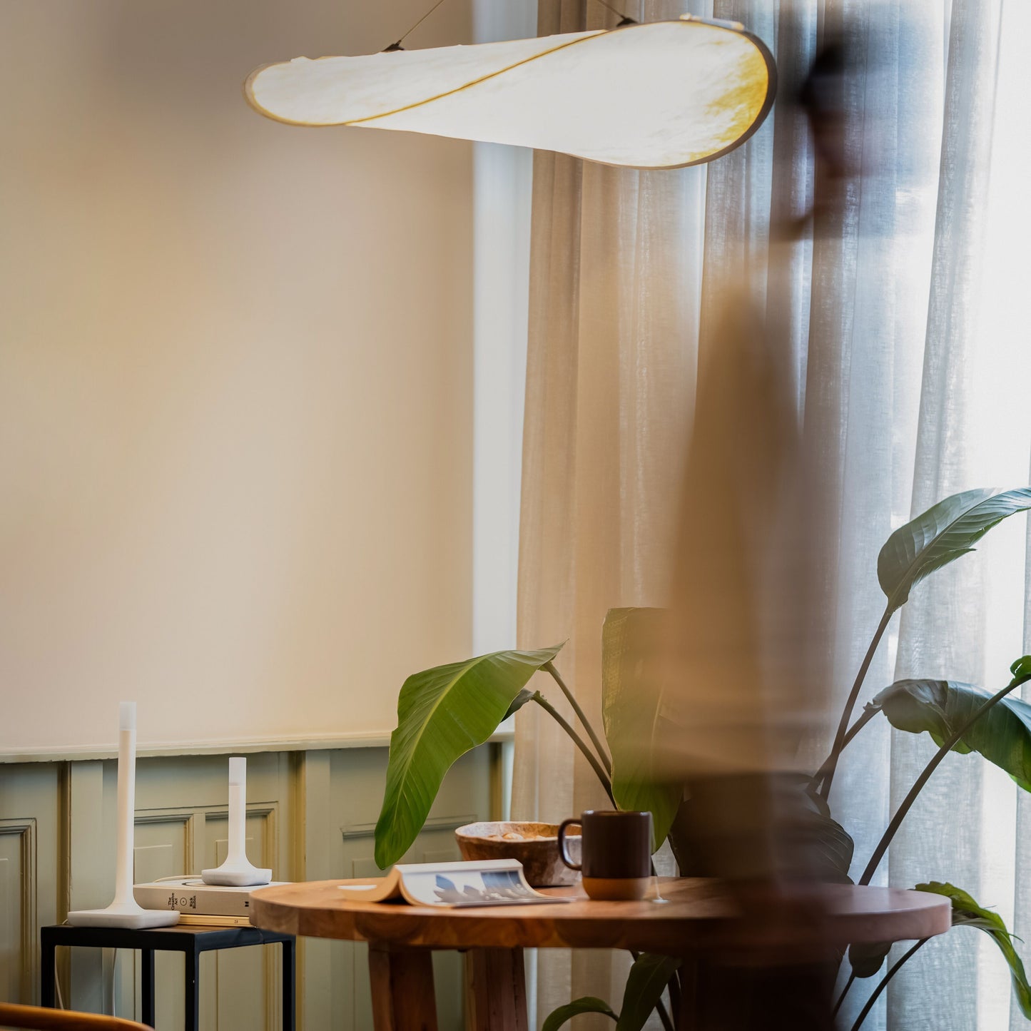 Home Assistant Connect ZBT-2 ved træbord med kaffekop, tallerken, bog og pendellampe over, lyse gardiner i baggrunden.