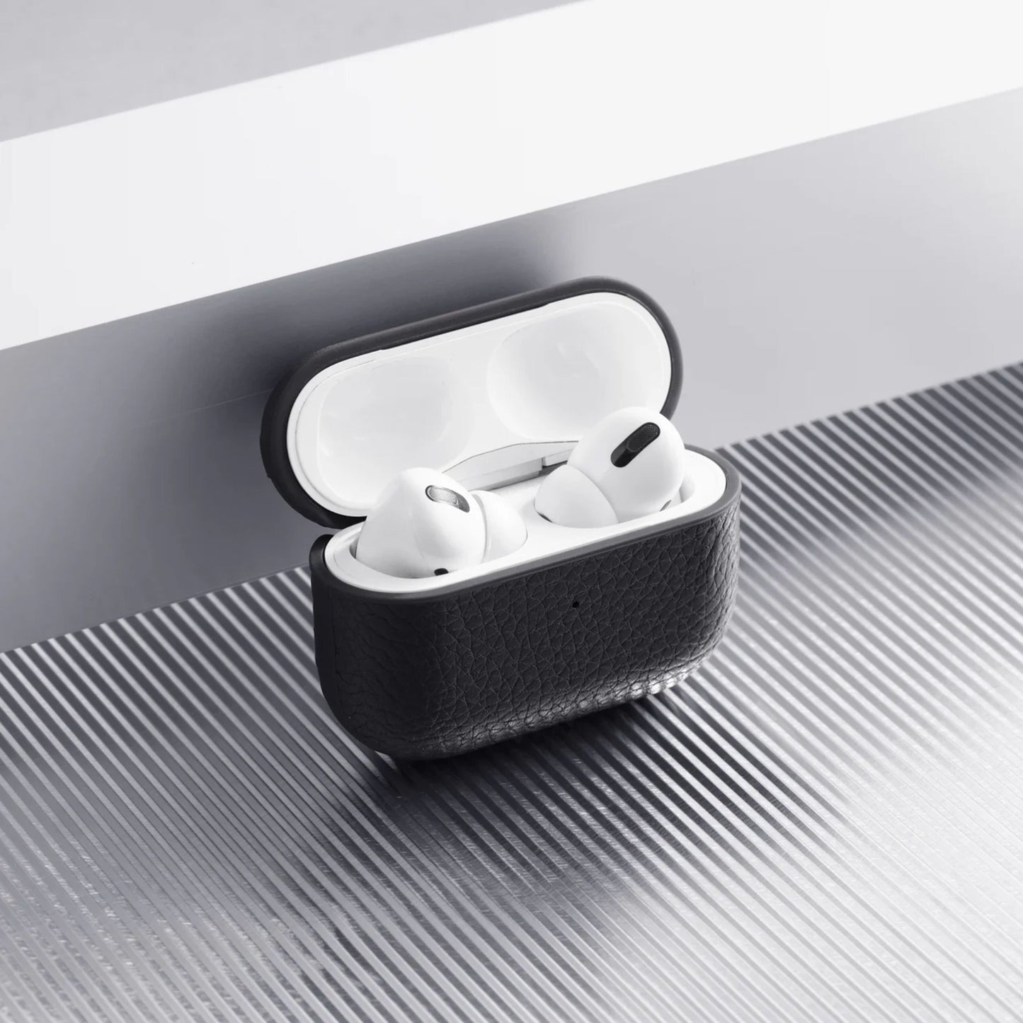 Apple AirPods Pro 3 etui i hvid i sort WOOLNUT læderetui på riflet metaloverflade ved hvidgrå kant.