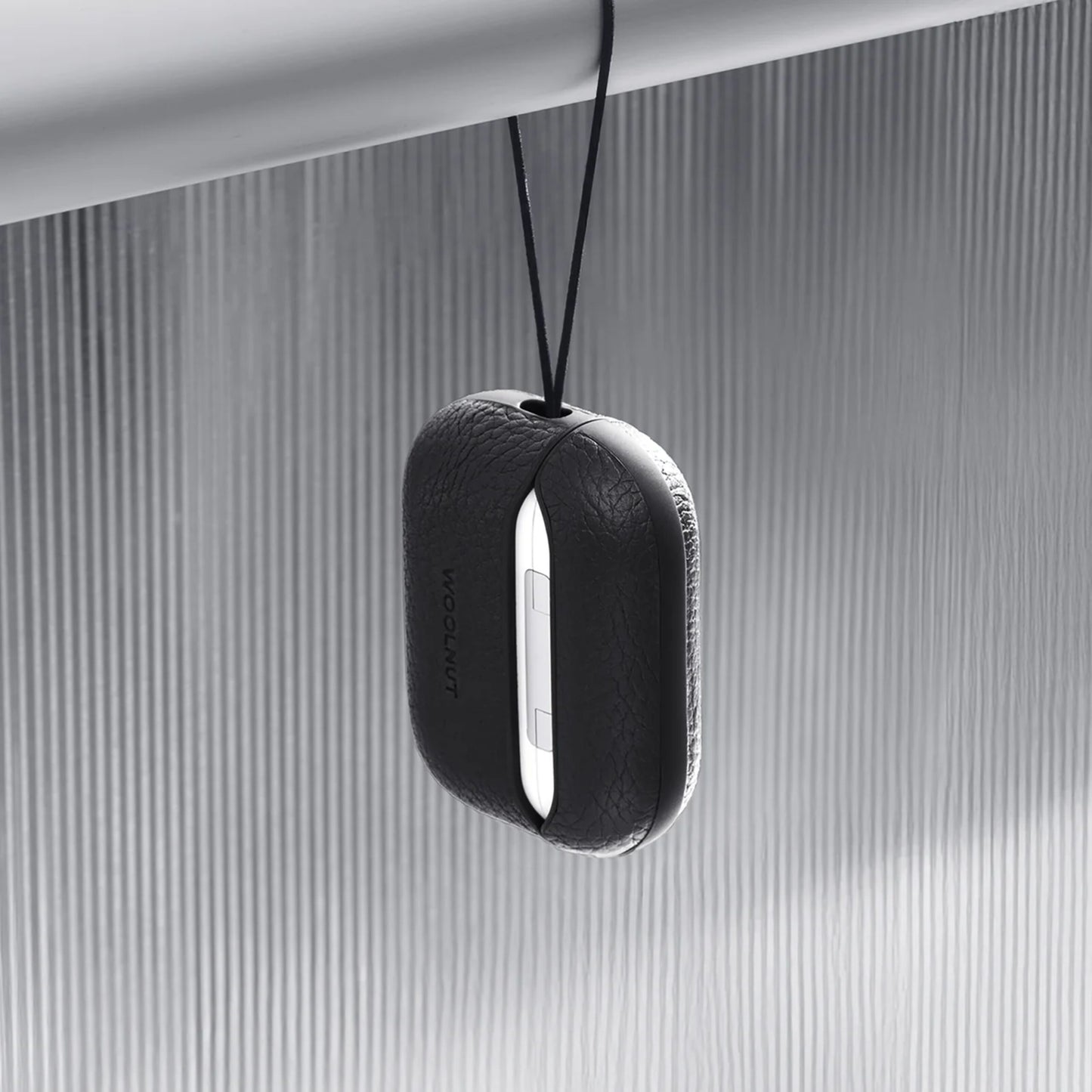 AirPods Pro 3 oplader i sort WOOLNUT læderetui hænger fra sort snor på metallisk stang, lodret rillet baggrund.