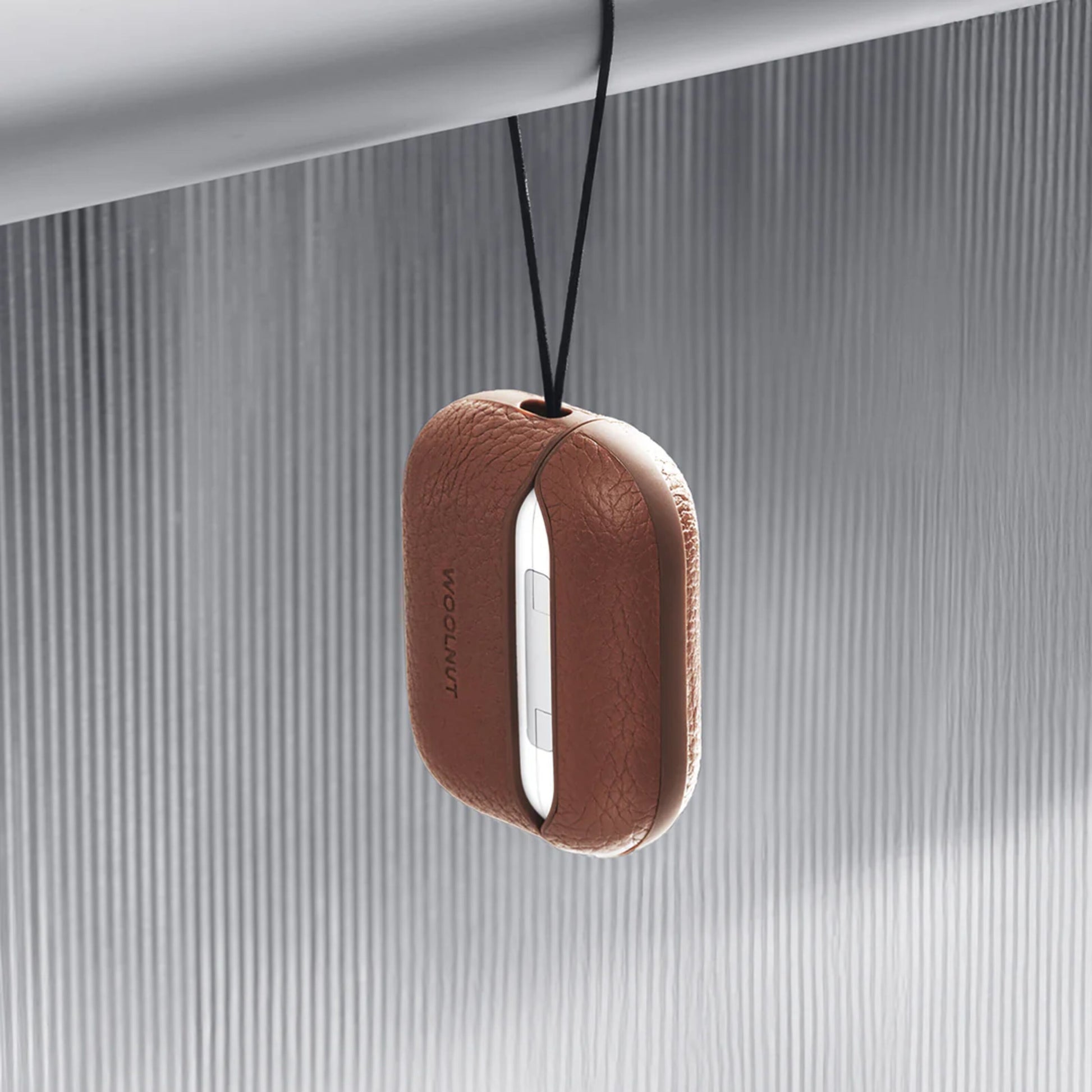 WOOLNUT læderetui til AirPods Pro 3 opladeretui, Cognac, hænger i sort snor fra metalstang foran tekstureret overflade.