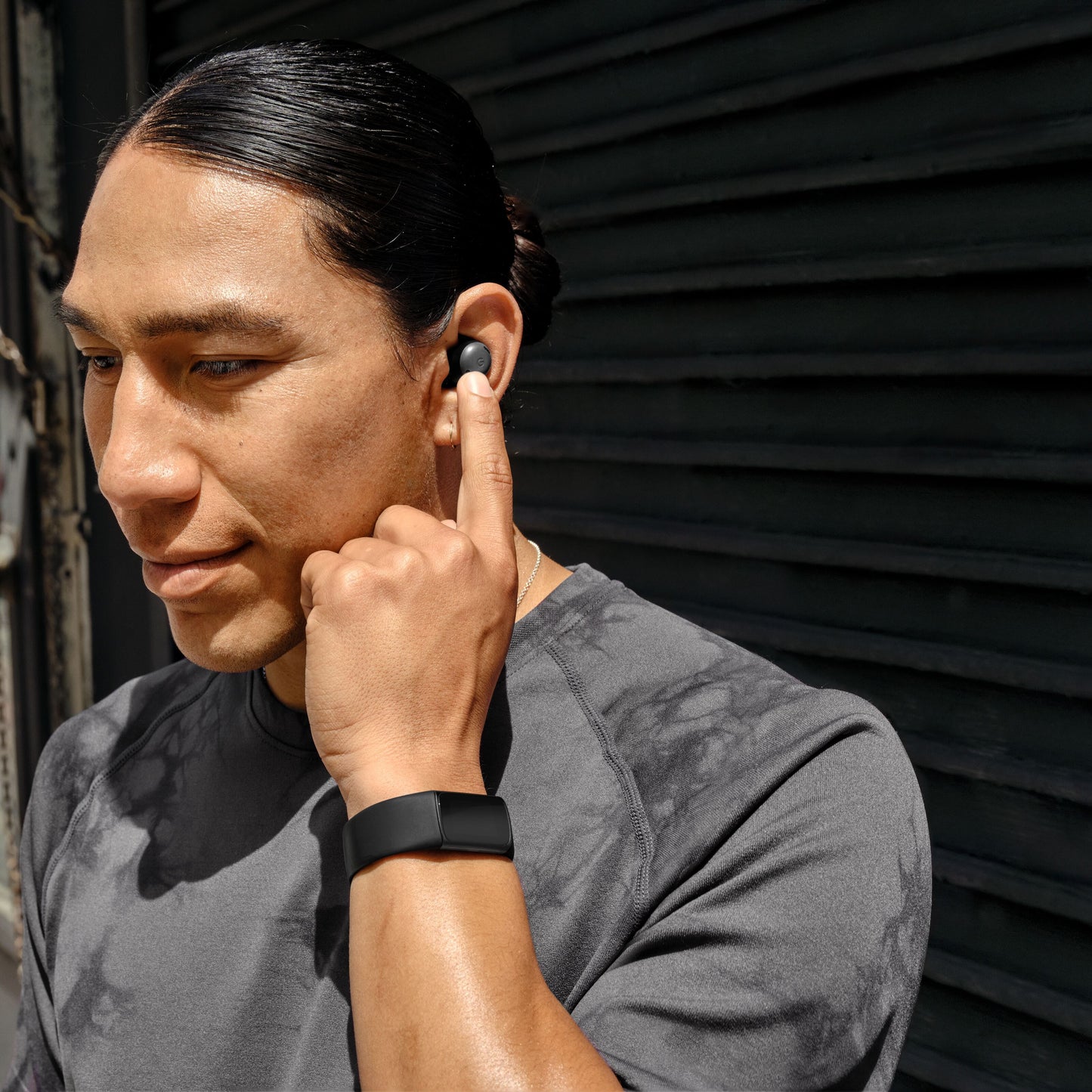 Fitbit Oplad 6 aktivitetsarmbånd på håndled og trådløs earbud i øret, person i grå t-shirt.