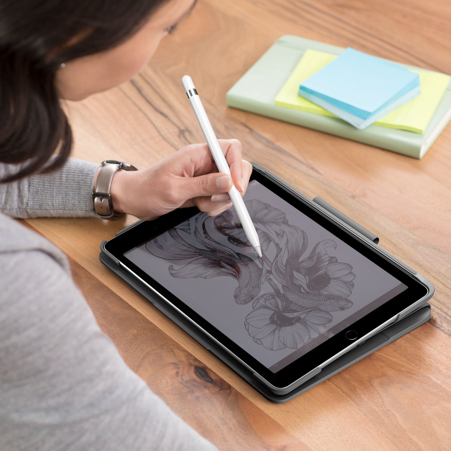 IPad 10,2 med Logitech Slim Folio tastaturcover og stylus bruges til at tegne, ses ved siden af lukket notesbog og sticky notes.