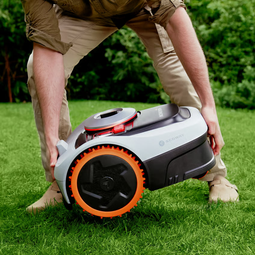 En person løfter en hvid og sort Segway Navimow i208E AWD robotplæneklipper med orange fælge hen over græsplænen.