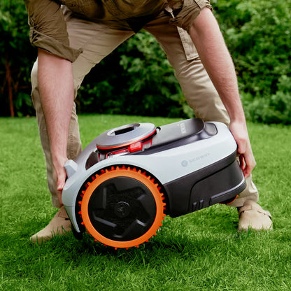 En person løfter en hvid og sort Segway Navimow i208E AWD robotplæneklipper med orange fælge hen over græsplænen.
