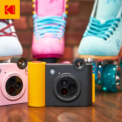 Kodak Smile+ digitalkamera i sort og lyserød med gule detaljer, ZINK-udskrivning og Bluetooth foran tre rulleskøjter.
