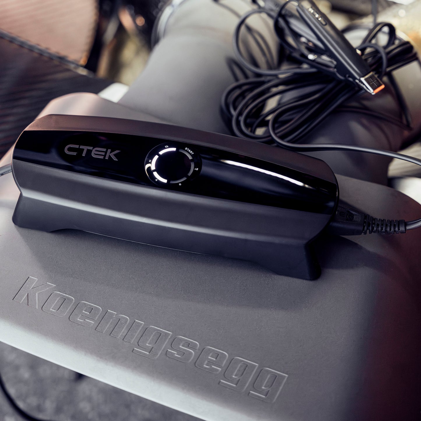 Ctek CS ONE Gen 2 12V batterioplader på mat overflade med Koenigsegg logo, kabler rullet sammen ved siden af.