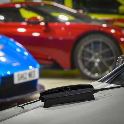 Ctek CS ONE Gen 2 sort smart batterioplader 12V på motorhjelm af hvid bil, uskarpe røde, blå og gule biler i baggrunden.