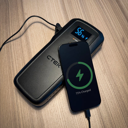 Ctek RB 3000 Startbooster 59,2 Wh sort med smartphone til opladning via kabel på lys træoverflade.
