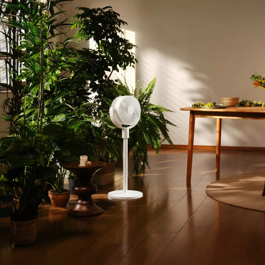 Xiaomi Smart Standing Air Circulation Fan i hvidt, placeret ved grønne planter på et trægulv tæt på et lavt træbord med skåle.