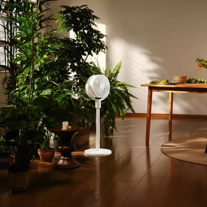 Xiaomi Smart Standing Air Circulation Fan i hvidt, placeret ved grønne planter på et trægulv tæt på et lavt træbord med skåle.