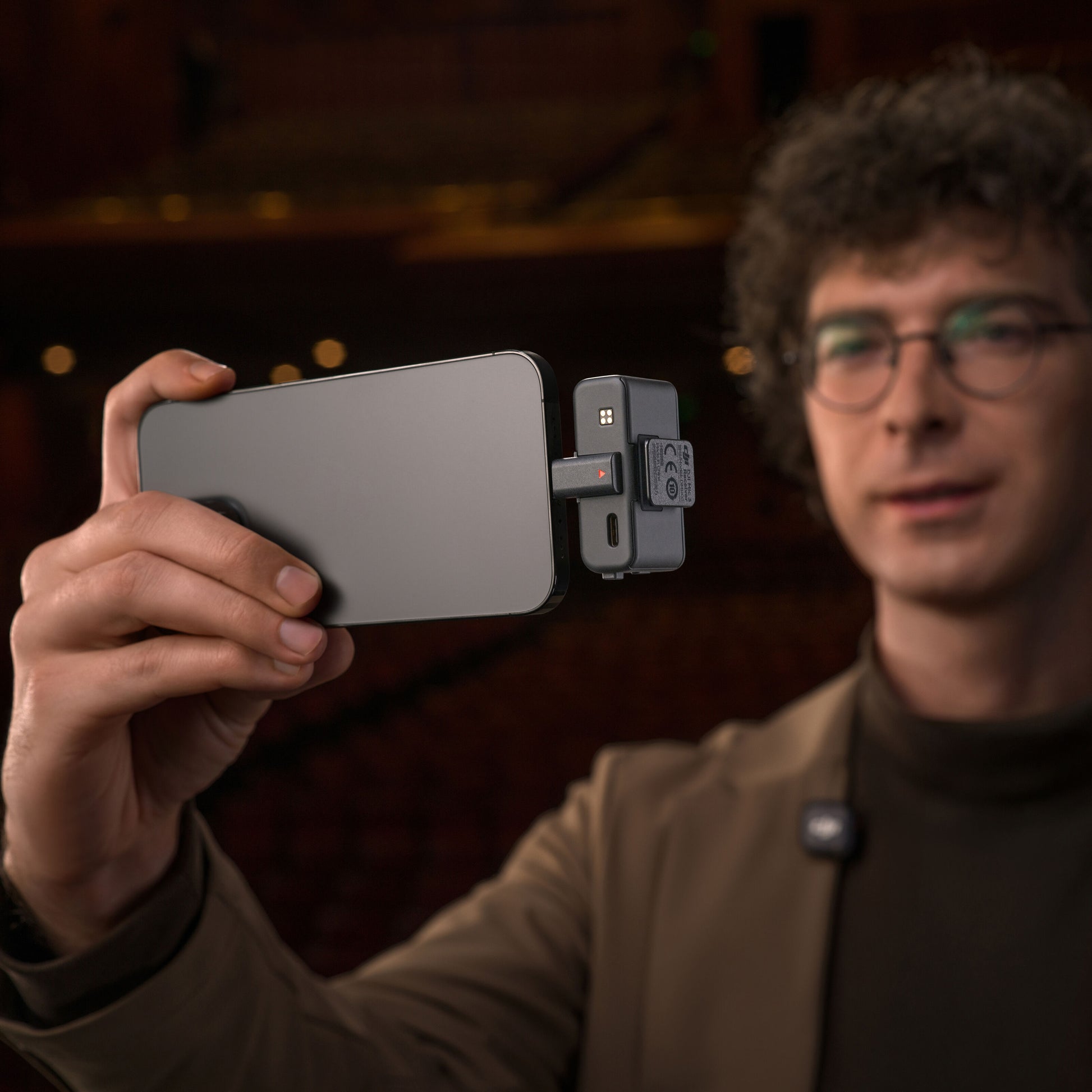 DJI Mic 3 trådløst mikrofonsystem 1 TX + 1 RX monteret på smartphone, holdt af person med krøllet hår og briller.