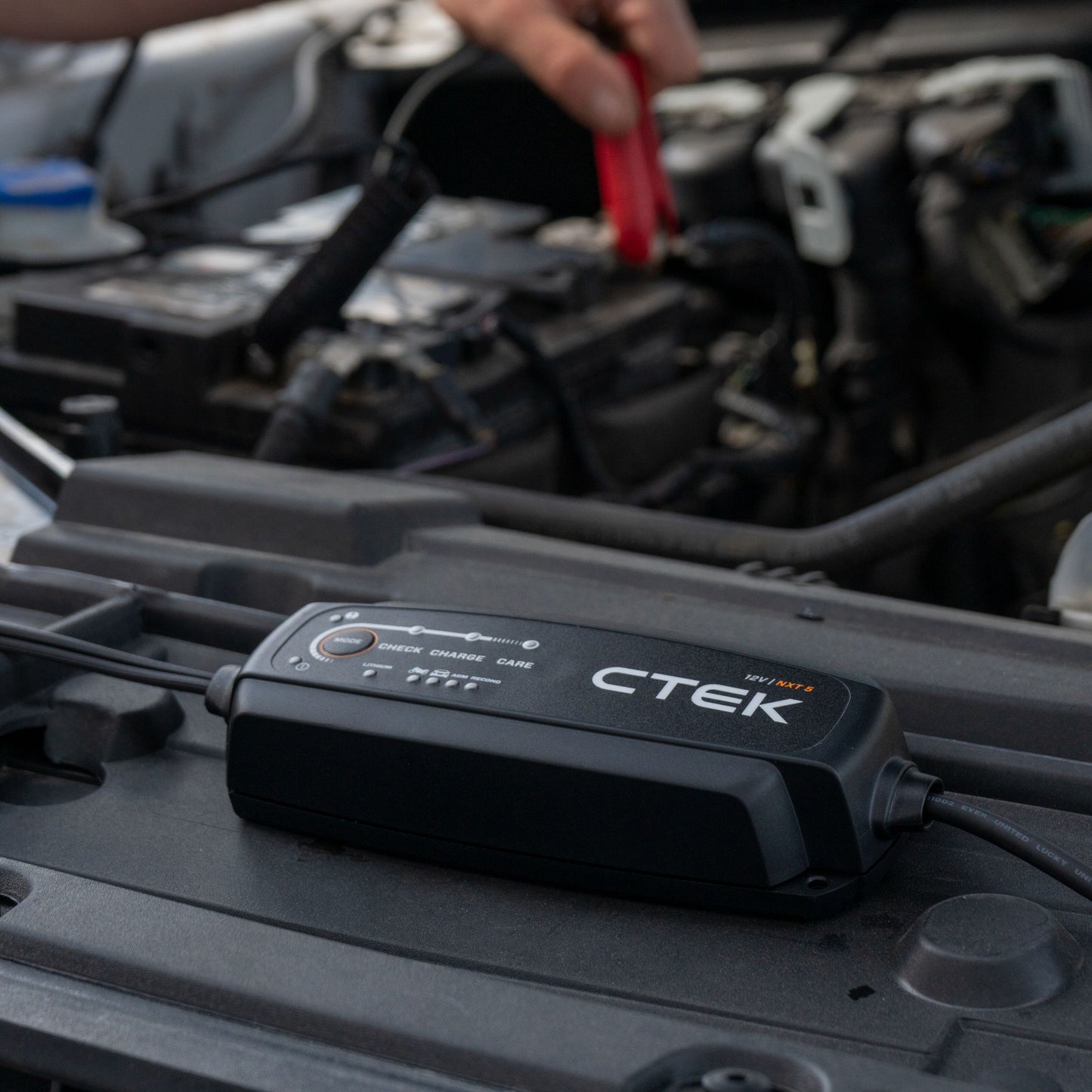 Ctek NXT 5 Smart batterioplader 12V 5A på bilmotor, rød klemme forbindes til batteri, motordele og kabler synlige.