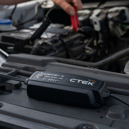 Ctek NXT 5 Smart batterioplader 12V 5A på bilmotor, rød klemme forbindes til batteri, motordele og kabler synlige.