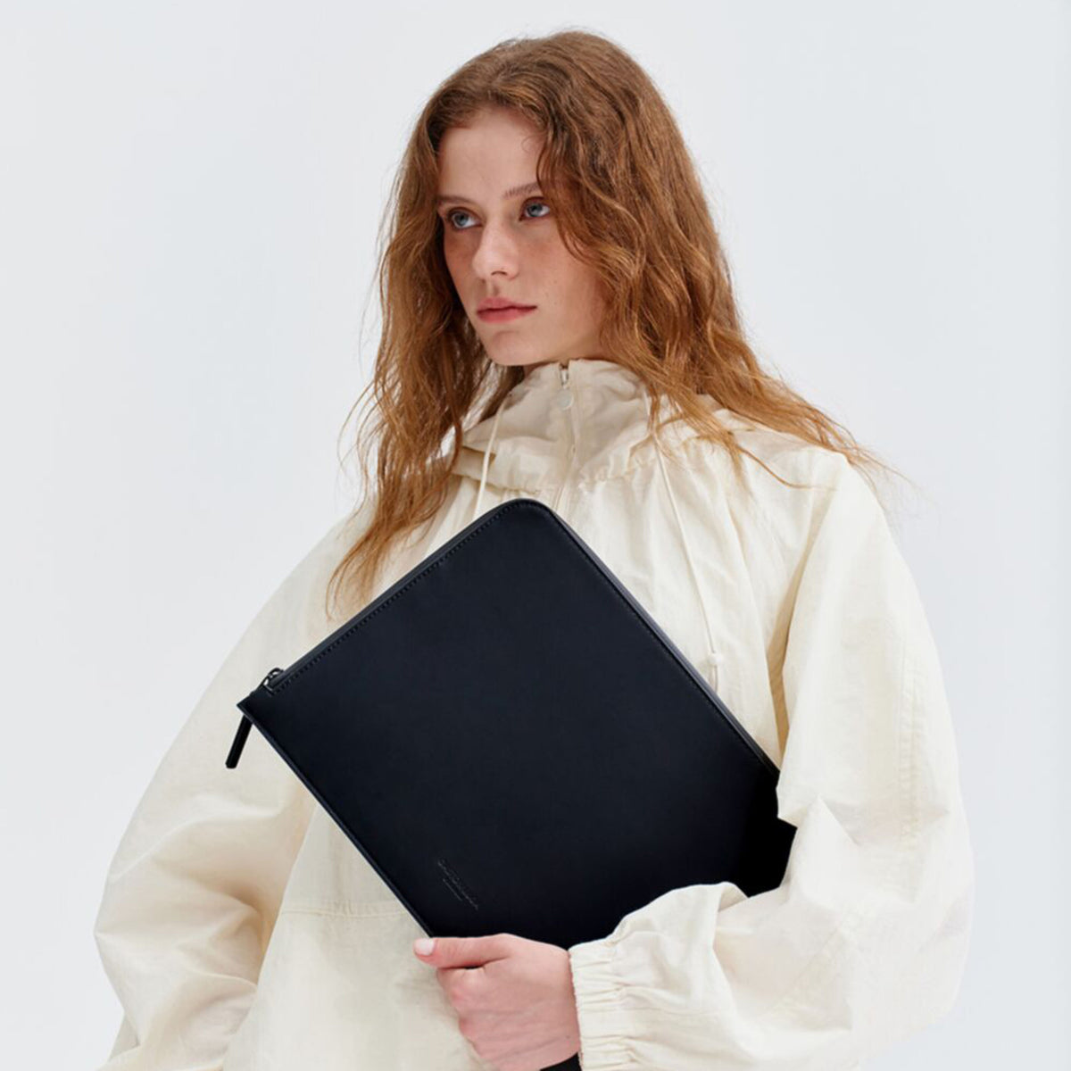 GASTON LUGA Däsh laptop-taske 13-14″, sort, genbrugt polyester, holdes af en person i en cremefarvet jakke.