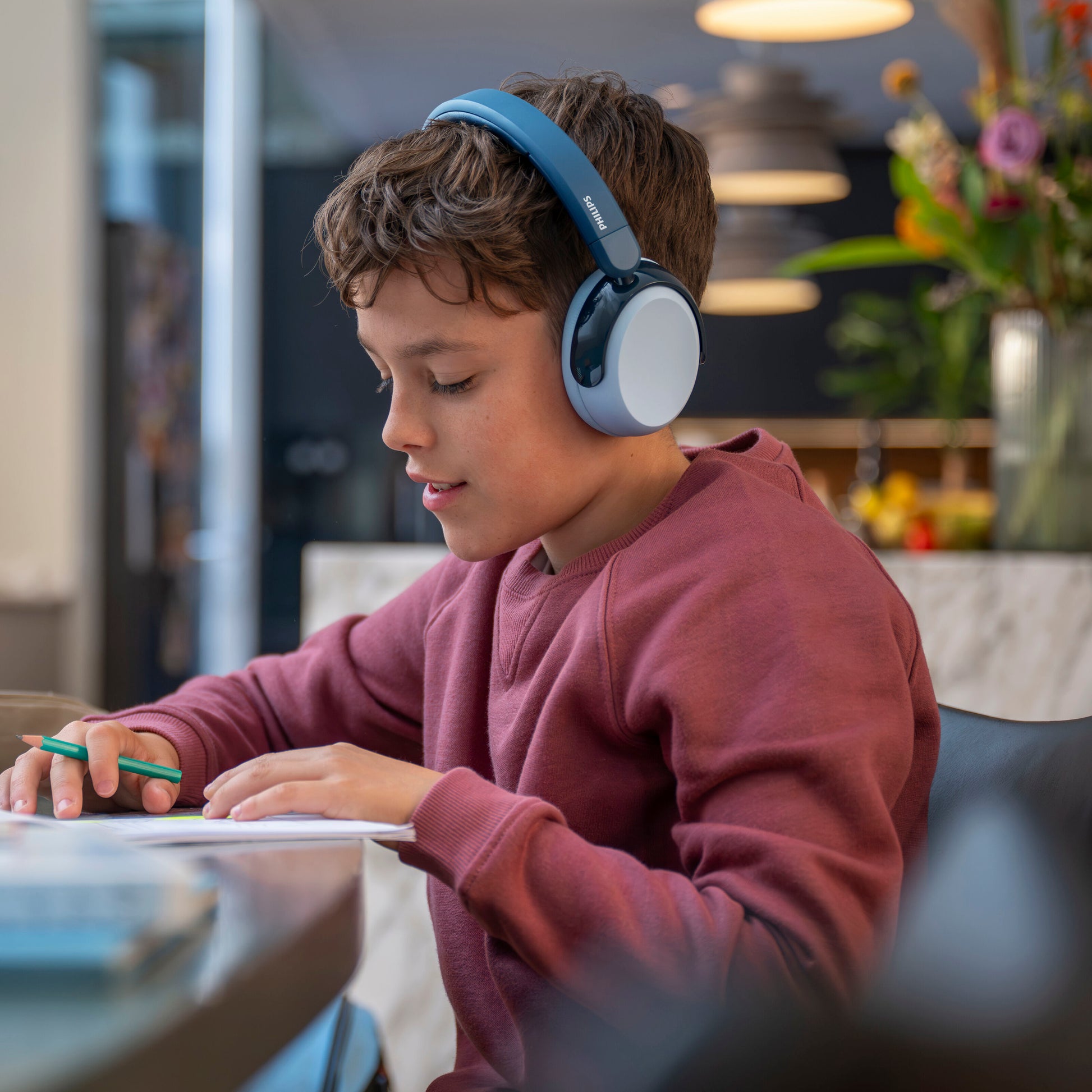 Dreng med Philips 5000-serien trådløse blå over-ear børnehøretelefoner, bærer sweatshirt, ved bord, skrivevinkel.