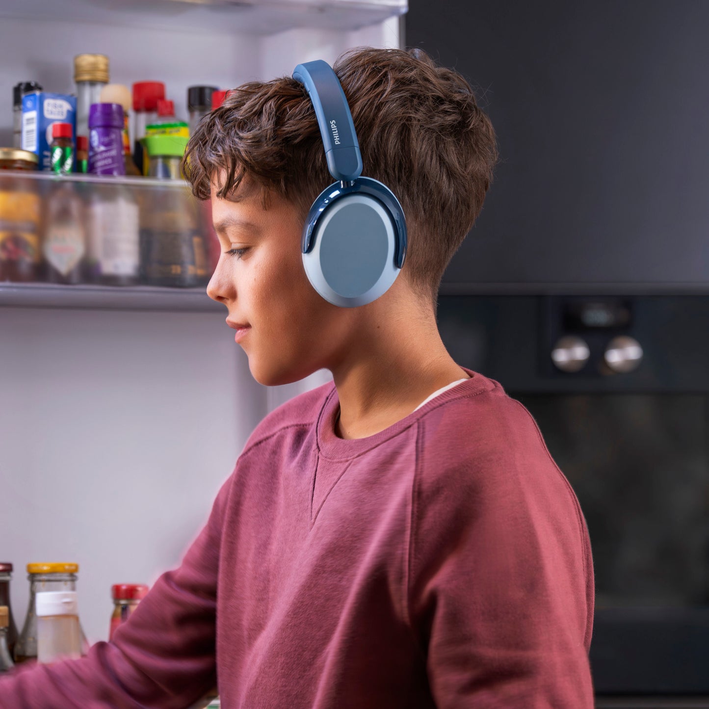Dreng med Philips 5000-serien trådløse over-ear børnehøretelefoner blå med volumenbegrænsning, ved åben køleskab.